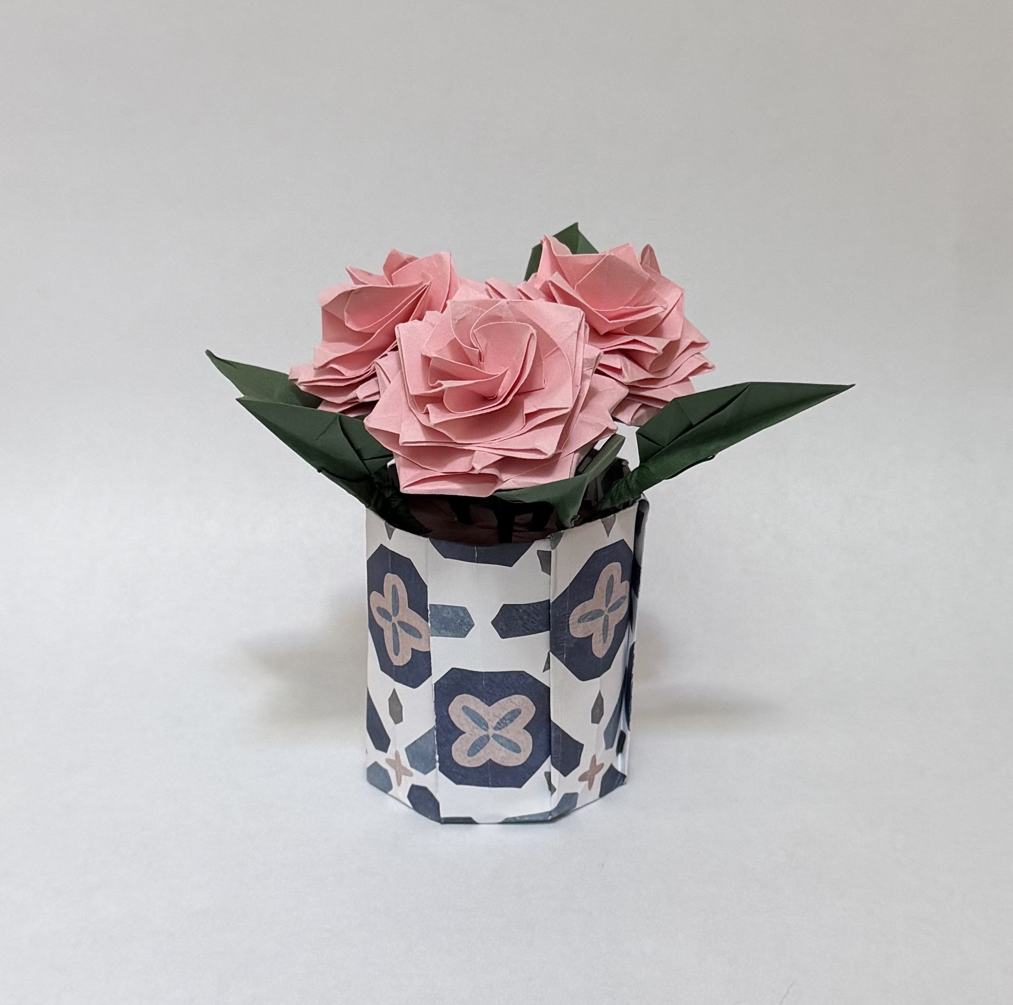 An image showing three pink roses in a blue and white pot with green leaves, created from paper.