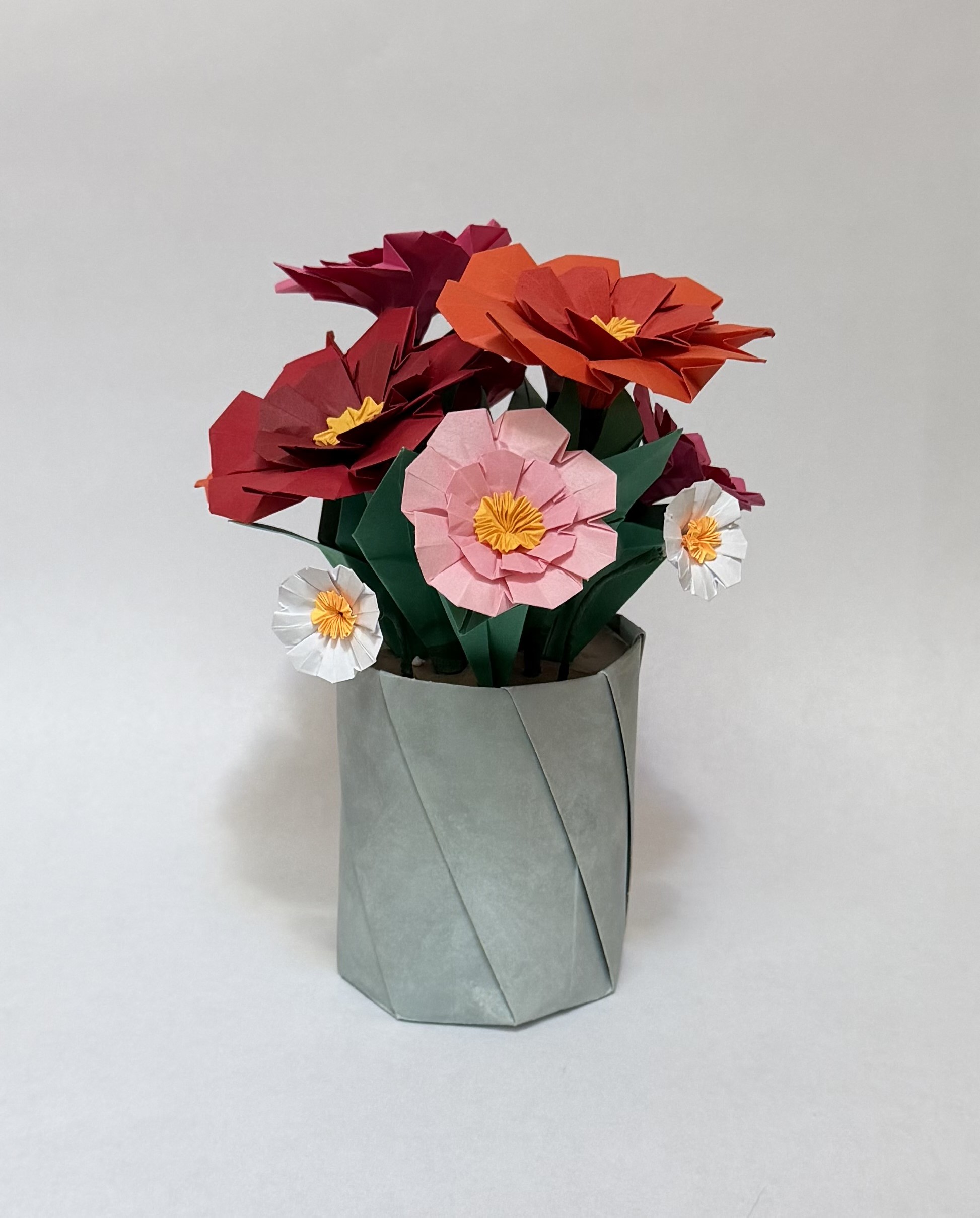 An image showing a collection of pink, orange, and red zinnias in a neutral pot with green leaves, created from paper.